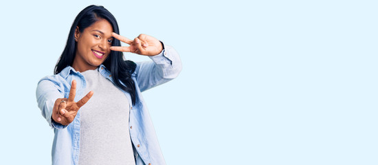 Hispanic woman with long hair wearing casual denim jacket smiling looking to the camera showing fingers doing victory sign. number two.