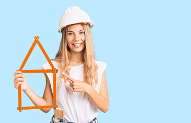 Beautiful caucasian woman with blonde hair wearing architect hardhat and holding tools smiling happy pointing with hand and finger