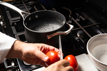 Black pot with water boiling on black stove with white bowl and red tomatoes being cut with knife by chef