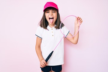 Young little girl with bang holding badminton racket sticking tongue out happy with funny expression.