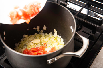 Black pot on stove with oil, onions and garlic adding tomatoes preparing a meal