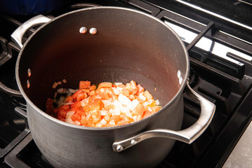 Black pot on stove with oil, onions and garlic adding tomatoes preparing a meal