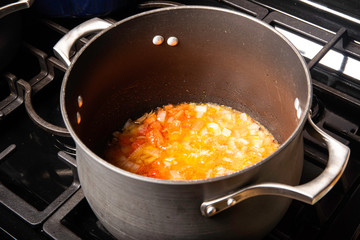 Black pot on stove with oil, onions and garlic adding tomatoes preparing a meal