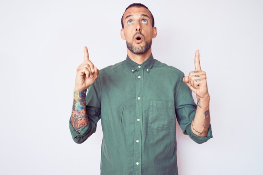 Young handsome man with tattoo wearing casual clothes amazed and surprised looking up and pointing with fingers and raised arms.