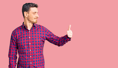 Handsome young man with bear wearing casual shirt looking proud, smiling doing thumbs up gesture to the side