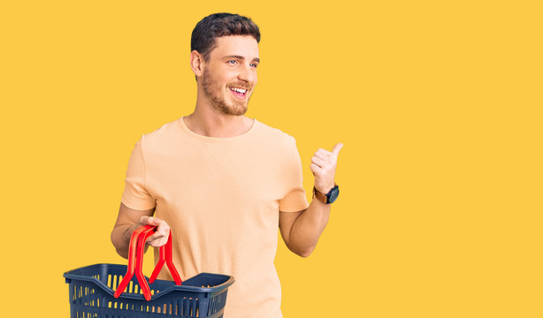 Handsome young man with bear holding supermarket shopping basket pointing thumb up to the side smiling happy with open mouth
