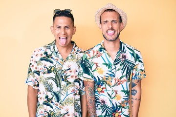 Young gay couple of two men wearing summer hat and hawaiian shirt sticking tongue out happy with funny expression. emotion concept.