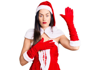 Young beautiful caucasian woman wearing santa claus costume swearing with hand on chest and open...