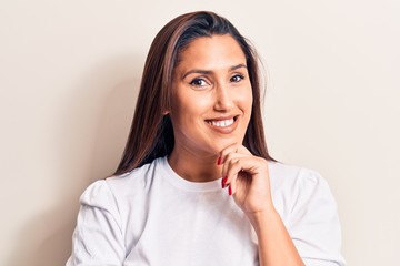 Young beautiful brunette woman wearing casual t-shirt smiling looking confident at the camera with crossed arms and hand on chin. thinking positive.