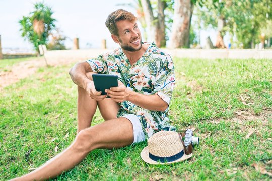 Caucasian handsome man smiling happy using touchpad device sitting on the grass at the park