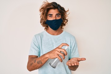 Young hispanic man wearing covid-19 mask using hand sanitizer gel making fish face with mouth and squinting eyes, crazy and comical.