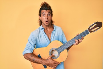 Young hispanic man playing classical guitar in shock face, looking skeptical and sarcastic, surprised with open mouth