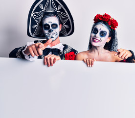 Young couple wearing mexican day of the dead costume holding blank empty banner pointing fingers to camera with happy and funny face. good energy and vibes.