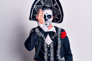 Young man wearing mexican day of the dead costume over white covering one eye with hand, confident smile on face and surprise emotion.