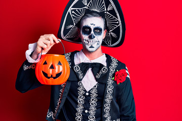 Young man wearing mexican day of the dead costume holding pumpkin looking positive and happy standing and smiling with a confident smile showing teeth