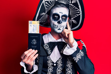 Young man wearing mexican day of the dead costume holding united states passport and boarding pass serious face thinking about question with hand on chin, thoughtful about confusing idea