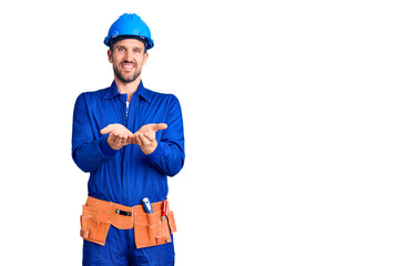 Young handsome man wearing worker uniform and hardhat smiling with hands palms together receiving or giving gesture. hold and protection