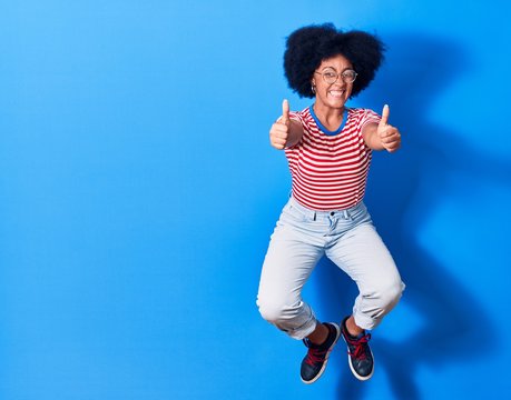 Young beautiful african american woman wearing casual clothes and glasses smiling happy. Jumping with smile on face doing ok sign with thumbs up over isolated blue background