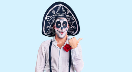 Man wearing day of the dead costume over background surprised pointing with hand finger to the side, open mouth amazed expression.
