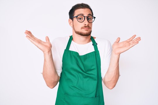 Young handsome man wearing apron clueless and confused with open arms, no idea and doubtful face.