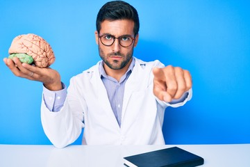 Young hispanic man sitting at the table wearing doctor coat holding brain pointing with finger to...