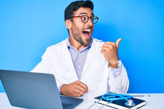 Young hispanic man wearing doctor uniform working at the clinic pointing thumb up to the side smiling happy with open mouth