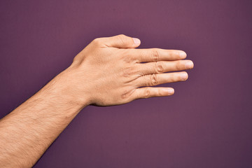 Hand of caucasian young man showing fingers over isolated purple background stretching and reaching with open hand for handshake, showing back of the hand