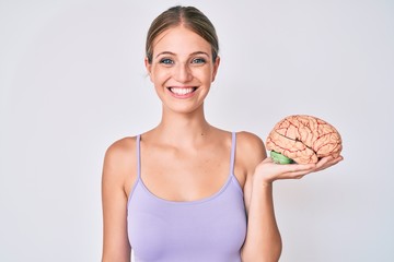 Young blonde girl holding brain looking positive and happy standing and smiling with a confident...