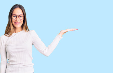 Beautiful brunette young woman wearing casual white sweater and glasses smiling cheerful presenting and pointing with palm of hand looking at the camera.