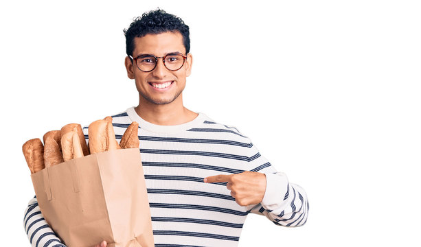 Hispanic handsome young man holding paper bag with bread pointing finger to one self smiling happy and proud