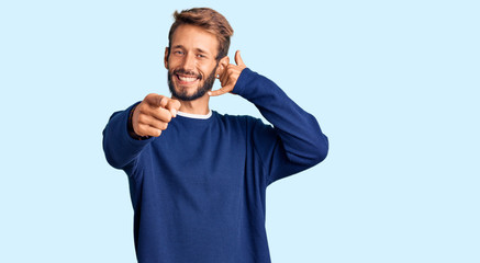 Handsome blond man with beard wearing casual sweater smiling doing talking on the telephone gesture and pointing to you. call me.