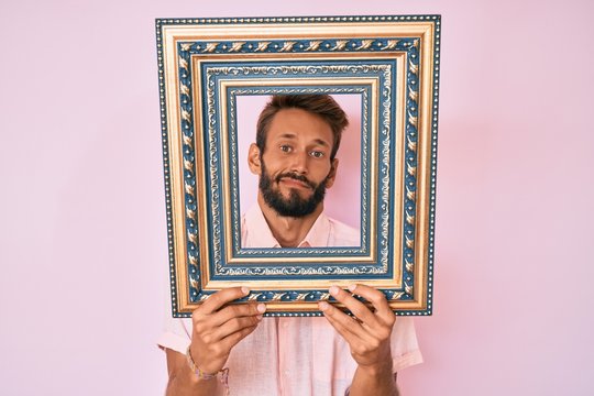 Handsome caucasian man with beard holding empty frame clueless and confused expression. doubt concept.