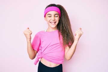 Obraz na płótnie Canvas Cute hispanic child girl wearing sportswear very happy and excited doing winner gesture with arms raised, smiling and screaming for success. celebration concept.