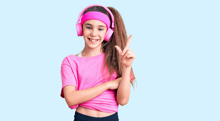 Obraz na płótnie Canvas Cute hispanic child girl wearing gym clothes and using headphones with a big smile on face, pointing with hand and finger to the side looking at the camera.