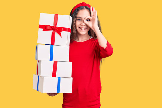Cute hispanic child girl holding gift smiling happy doing ok sign with hand on eye looking through fingers
