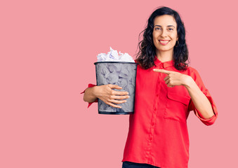 Young beautiful hispanic woman holding paper bin full of crumpled papers smiling happy pointing with hand and finger