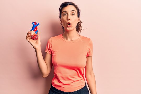Young beautiful hispanic woman wearing sportswear holding heart scared and amazed with open mouth for surprise, disbelief face