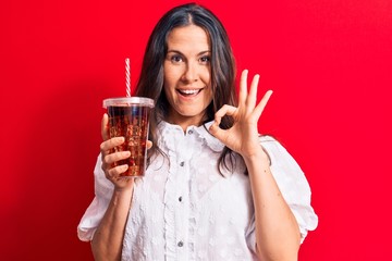 Beautiful brunette woman drinking cola refreshment beverage using straw over red background doing ok sign with fingers, smiling friendly gesturing excellent symbol