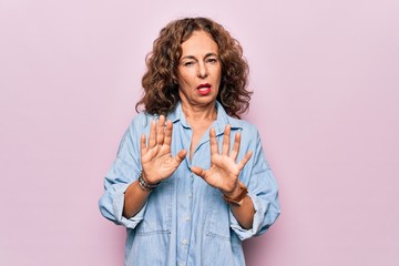 Middle age beautiful woman wearing casual denim shirt standing over pink background Moving away...