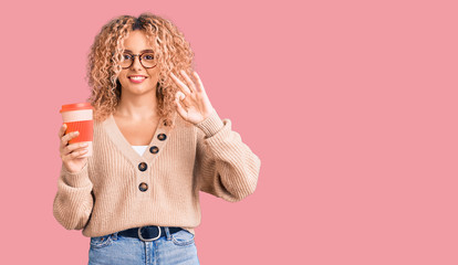 Young blonde woman with curly hair wearing glasses and drinking a cup of coffee doing ok sign with fingers, smiling friendly gesturing excellent symbol