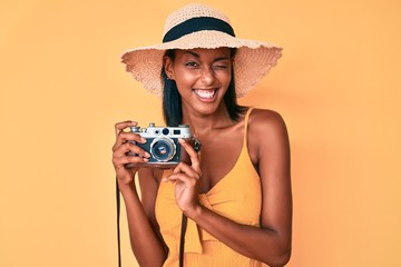 Young african american woman wearing summer hat holding vintage camera winking looking at the camera with sexy expression, cheerful and happy face.