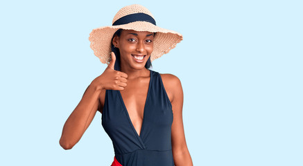 Young african american woman wearing swimsuit and summer hat doing happy thumbs up gesture with hand. approving expression looking at the camera showing success.