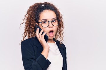 Beautiful kid girl with curly hair wearing glasses having conversation talking on the smartphone scared and amazed with open mouth for surprise, disbelief face