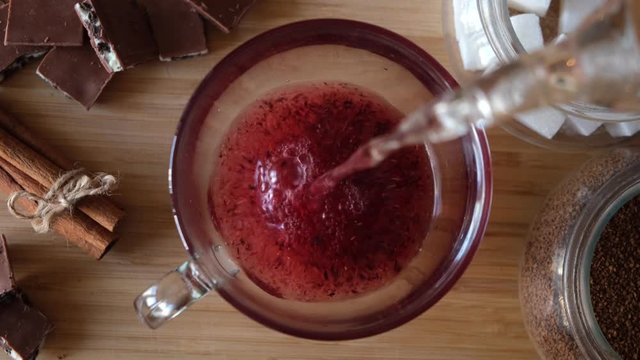 Process of brewing red karkade tea, close up top view. Concept. Cup of freshly brewed fruit tea, pouring hot water from the kettle into a glass cup on wooden background.