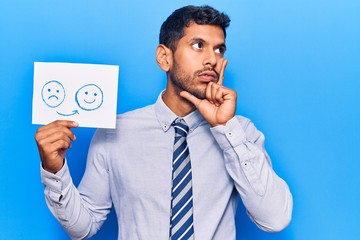 Young latin man holding sad to happy emotion paper serious face thinking about question with hand...