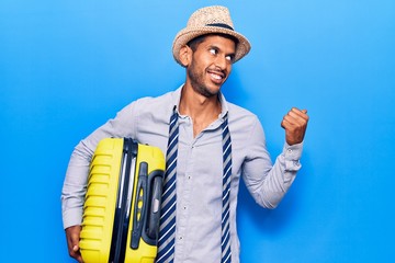 Young latin man wearing summer hat holding cabin bag pointing thumb up to the side smiling happy with open mouth