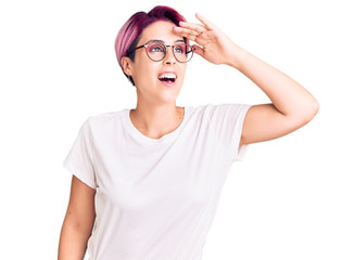 Young beautiful woman with pink hair wearing casual clothes and glasses very happy and smiling looking far away with hand over head. searching concept.