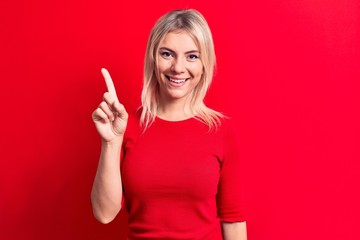 Young beautiful blonde woman wearing casual red t-shirt standing over isolated background smiling with an idea or question pointing finger up with happy face, number one