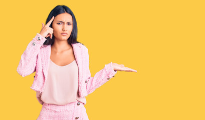 Young beautiful latin girl wearing business clothes confused and annoyed with open palm showing copy space and pointing finger to forehead. think about it.