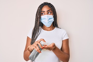 Young african american woman with braids wearing medical mask using hand sanitizer smiling looking to the side and staring away thinking.